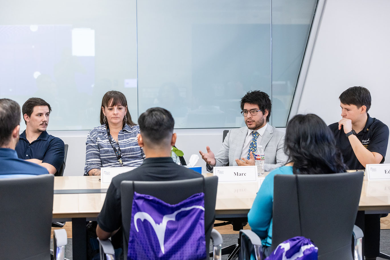 A male college student speaks to his peers while they are seated around a conference table.