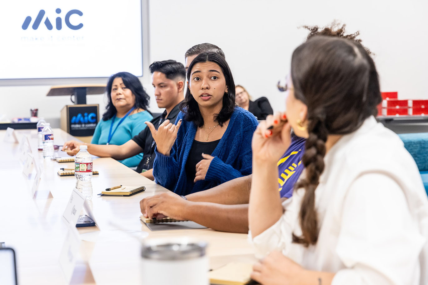 A female college student speaks to her peers while they are seated around a conference table.