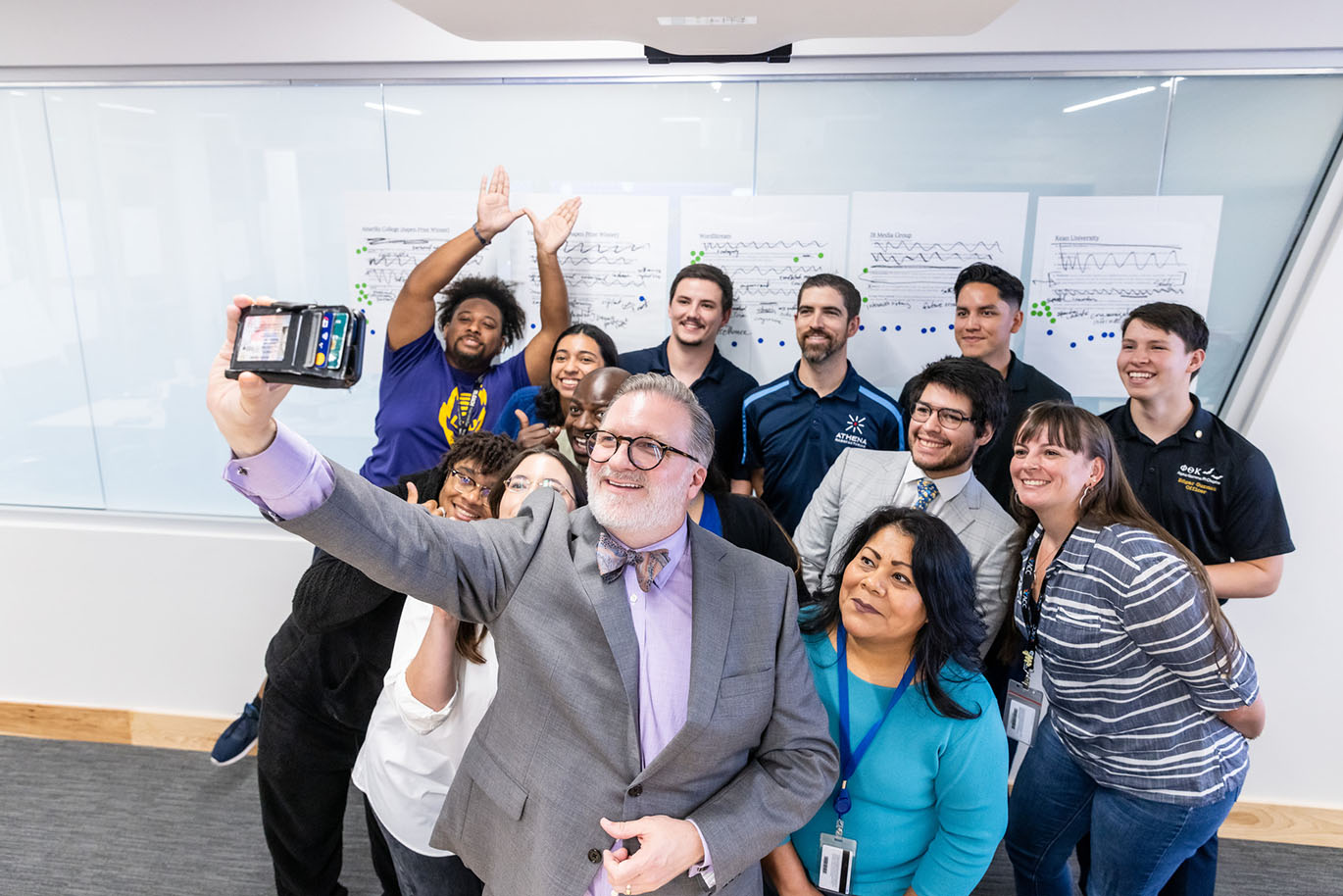 ACC Chancellor Dr. Russell Lowery-Hart takes a group selfie with a diverse group of college students representing various ages, genders, ethnicities, and races. Everyone is smiling. Behind them, a glass wall displays four sheets of white paper covered in printed text, handwritten black marker notes, and colorful round stickers.