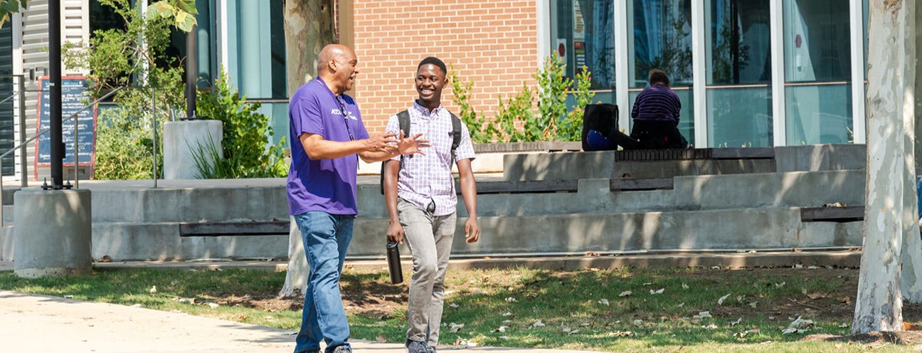 Two men in purple shirts engaged in conversation while standing on a sidewalk.