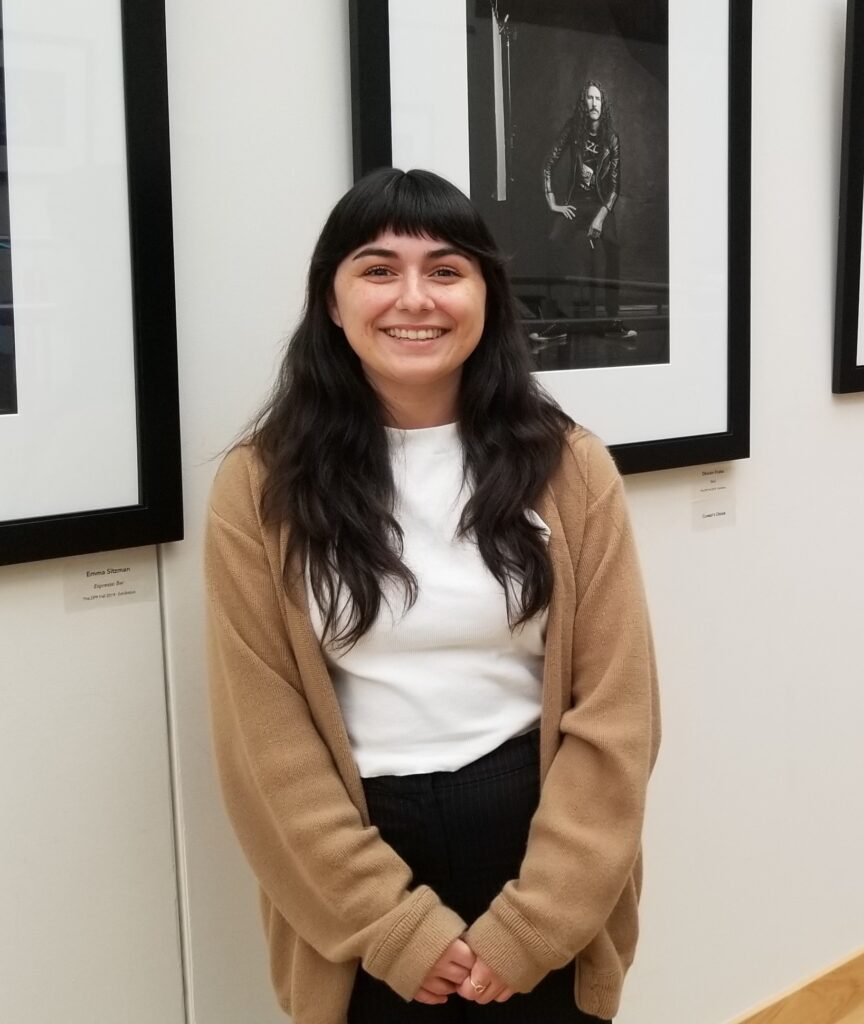 Alicia T. a former Peer Money Mentor is posing next to some framed photographs that are posted on the wall.