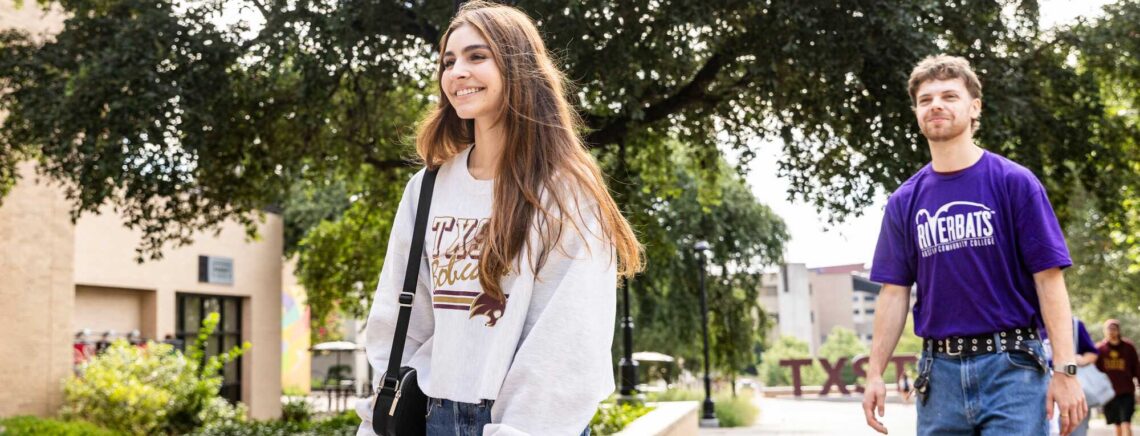 A female Texas State University student strolls along a sidewalk at Texas State University beside a man wearing a purple ACC Riverbats shirt.