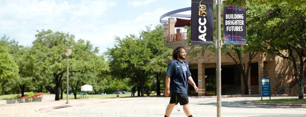 An ACC student walks on the sidewalk beside a sign that reads "ACC Building Brighter Futures", surrounded by greenery and urban scenery.