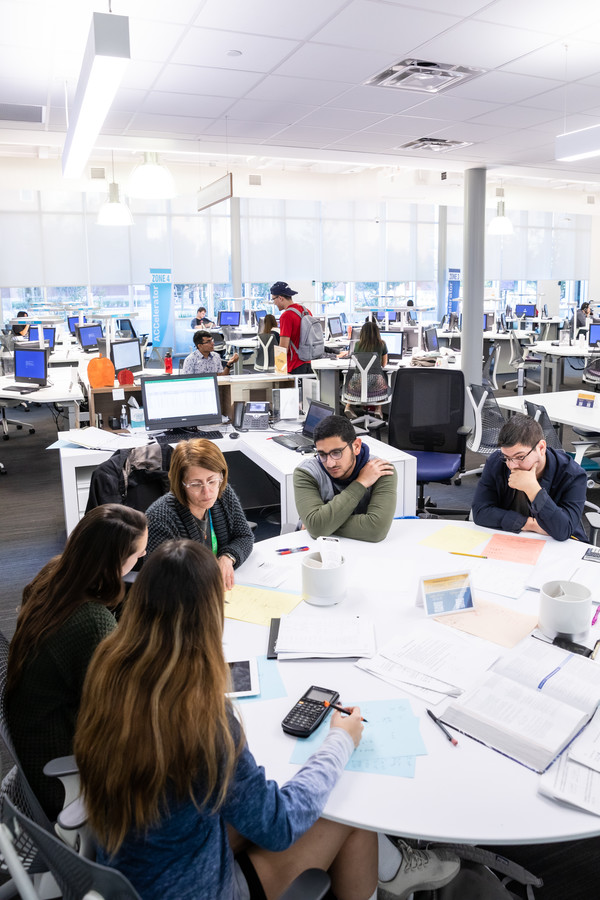 A group of six students is gathered around a large round table in a modern study space. They are engaged in discussion, with books, papers, and a calculator spread out on the table. In the background, several other students are working at individual desks with computers. The room is bright and spacious, featuring large windows that let in natural light.
