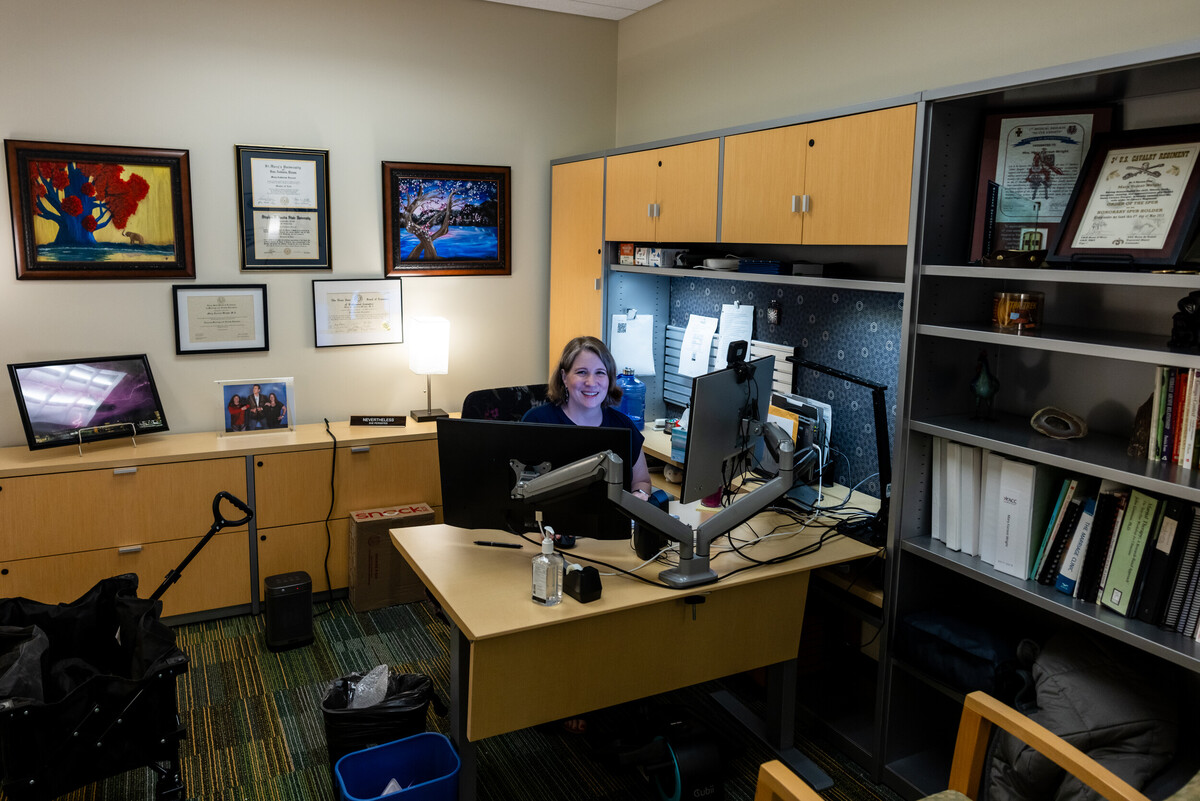 A well-organized office space featuring a woman sitting at a desk with dual computer monitors. The desk is equipped with various office supplies and personal items. Behind her, there are framed certificates and artwork on the wall. The room has a warm ambiance with a lamp providing light, and a bookshelf filled with documents and books is visible in the background. The floor has a patterned carpet, adding a touch of color to the space.