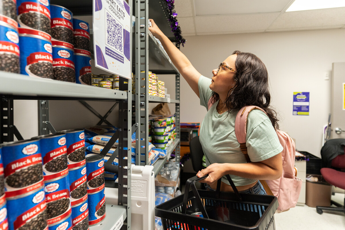A young woman with long dark hair and glasses is reaching for a can of black beans on a shelf in a food pantry. She is wearing a light green striped shirt and has a pink backpack slung over one shoulder. In her other hand, she holds a shopping basket. The shelves are stocked with various canned goods and food items, and the room has a welcoming atmosphere.