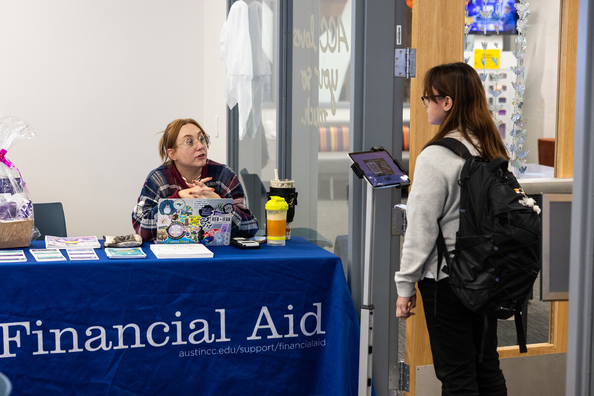 A woman with glasses and red hair, wearing a plaid shirt, is seated at a table covered with a blue tablecloth that reads "Financial Aid." She is engaged in conversation with another woman who has long brown hair and is wearing a backpack. The second woman is standing and looking at a tablet. In the background, there are various informational materials and decorations visible on the walls.