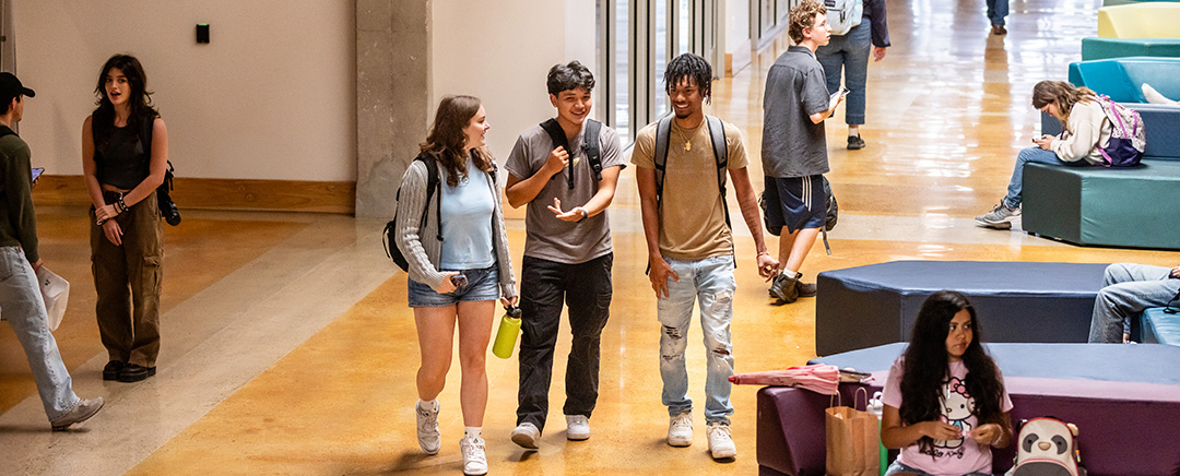 A busy indoor space in a university or college setting, featuring a mix of students walking and socializing. In the foreground, three students are chatting and walking together, while others are scattered throughout the area, some sitting on colorful couches and others moving through the space. The environment is bright and modern, with large windows allowing natural light to fill the room.